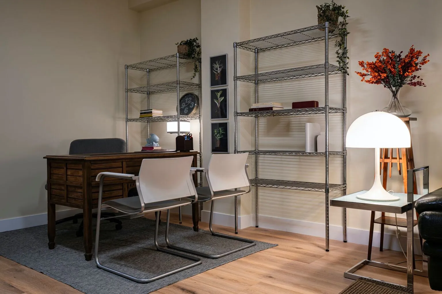 Oficina moderna con escritorio de madera, sillas blancas y estanterías metálicas. La decoración incluye plantas, libros y una lámpara de diseño, creando un ambiente acogedor y funcional.