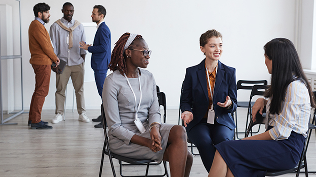 Grupo diverso de profesionales conversando en un entorno de oficina moderna, con dos mujeres sentadas en primer plano y hombres en el fondo, todos vestidos formalmente.