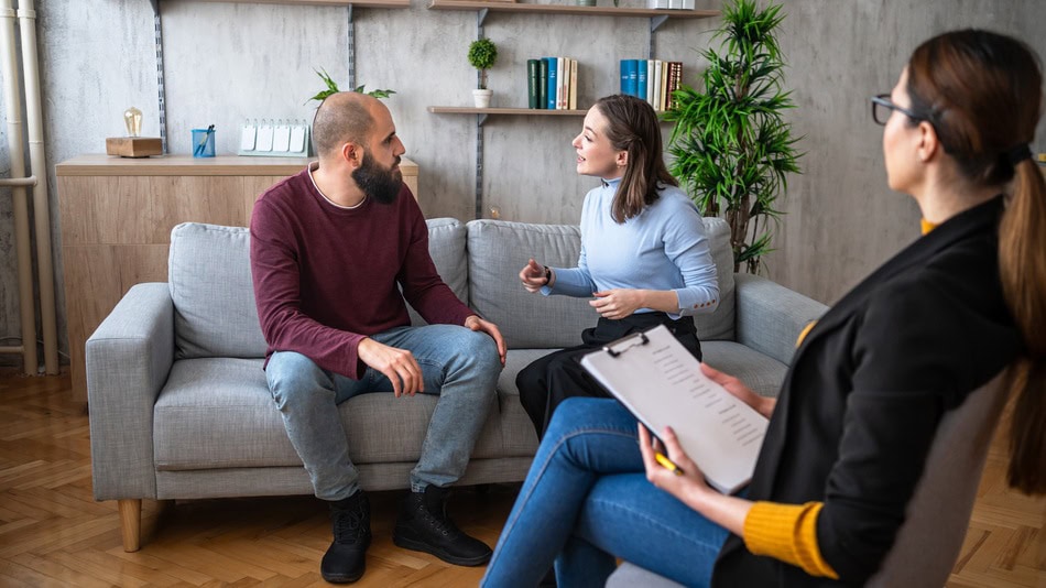 Pareja en sesión de terapia conversando con una terapeuta en un ambiente acogedor, con muebles modernos y estanterías de libros al fondo. La mujer habla expresivamente mientras el hombre escucha atentamente, y la terapeuta toma notas.
