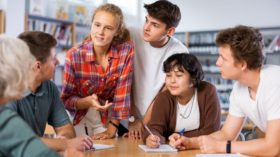 Grupo de estudiantes colaborando en una discusión en una biblioteca, con una joven liderando la conversación mientras los demás toman notas y escuchan atentamente.