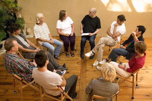 Grupo de personas en una sesión de terapia grupal, sentadas en círculo en un ambiente acogedor, con un facilitador hablando y los participantes escuchando atentamente. La imagen muestra un entorno natural con luz suave, ideal para la reflexión y el intercambio emocional.