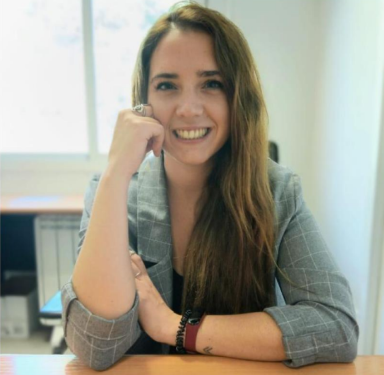 Mujer sonriente con cabello largo y castaño, vestida con un blazer gris, sentada en un escritorio de oficina. La imagen muestra un ambiente de trabajo luminoso y profesional.